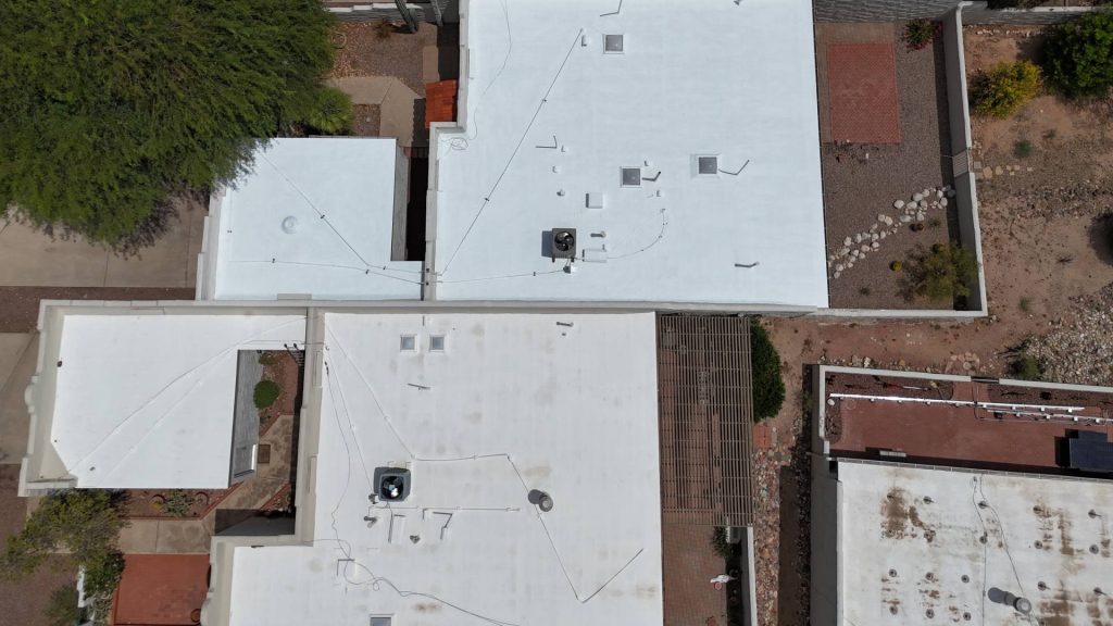 Aerial view of white, flat-roofed buildings with HVAC units and wiring on top. The surrounding area includes greenery, patios, stone paths, and a fenced yard with gravel and scattered plants.