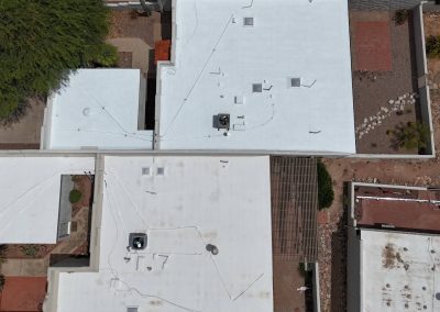 Aerial view of white, flat-roofed buildings with HVAC units and wiring on top. The surrounding area includes greenery, patios, stone paths, and a fenced yard with gravel and scattered plants.