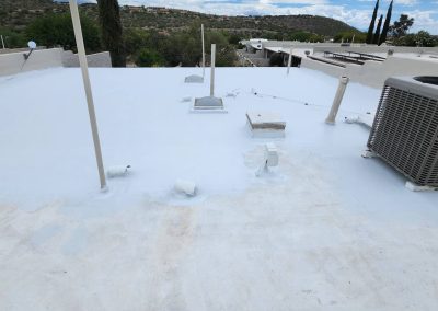 A flat white rooftop with HVAC unit, pipes, and small vents; distant houses and green hills are visible under a partly cloudy sky.