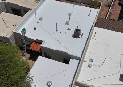 Aerial view of a flat white rooftop with various vents, pipes, and an air conditioning unit. A ladder is positioned against the side of the building near a red awning above a doorway. Adjacent rooftops are also visible.