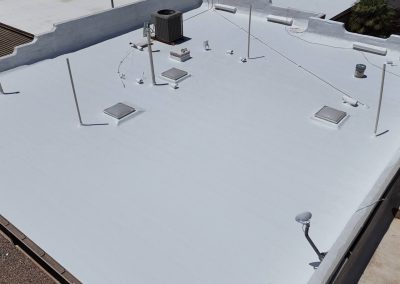 A person applies white coating to a flat rooftop with various vents, skylights, and equipment, creating a smooth, reflective surface under daylight.