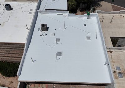 Aerial view of a flat white roof with various vents, pipes, and equipment, surrounded by neighboring roofs and a small courtyard area with plants and pavement.