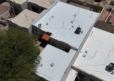A person on a ladder applies white coating to a flat roof of a building, surrounded by other rooftops, trees, and a gravel yard in a residential area.
