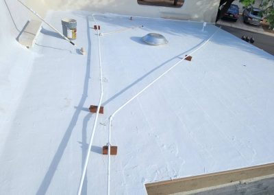 A flat, white rooftop with several white cables or pipes secured by brackets, a metal vent or dome in the center, and a paint bucket near the wall. Trees and parked cars are visible in the background.
