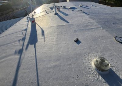 A white, flat rooftop in sunlight with various vents, pipes, and equipment casting long shadows; a small round dome and a cylindrical object are visible, along with trees and neighboring roofs in the background.