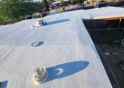 A flat rooftop coated in white sealant with paint cans and tools near the edge; a street, cars, and trees are visible in the background. Shadows from rooftop vents and objects are cast on the surface.