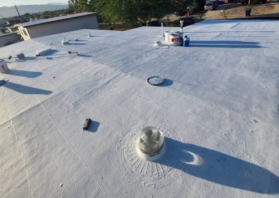 A flat white rooftop with vents, pipes, paint cans, and tools scattered around, under bright sunlight. Nearby houses, trees, and parked cars are visible in the background.