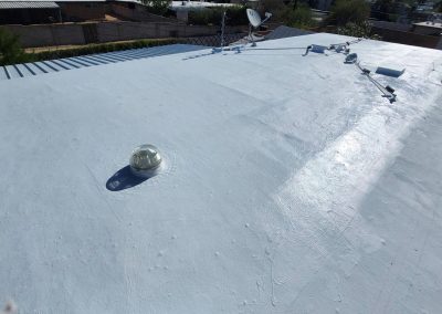 A flat, white-painted rooftop with a small dome-shaped skylight and several antennas, surrounded by neighboring buildings and greenery in the background.