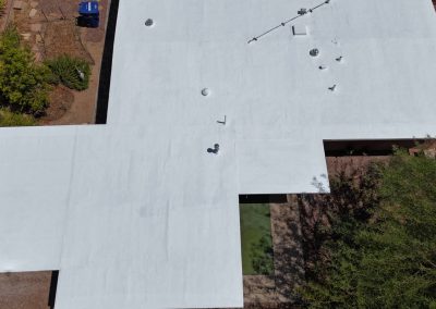 Aerial view of a flat, white roof on a house surrounded by dry landscaping, trees, and a small courtyard area. Several vents and pipes are visible on the roof.