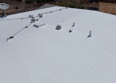 A flat white rooftop with several small vents, a satellite dish, and metal cables, seen from above, with neighboring yards and fences visible in the background.