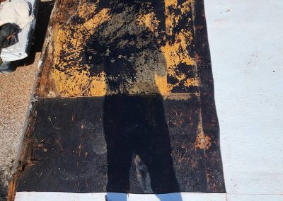 A person’s shadow is cast onto a partially stripped black and yellow roof surface, with a painted white section on the right. A vehicle with supplies is parked on the left edge of the photo.