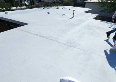 A person stands on a flat white roof, holding a bucket and roller, likely applying a coating. Several pipes and vents protrude from the roof under a clear sky, with nearby buildings visible in the background.