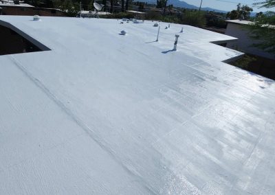 A wide view of a flat rooftop covered in a smooth, white reflective coating, with several vent pipes and nearby buildings visible in the background under a clear sky.