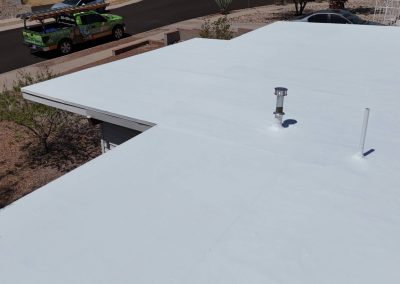 A flat, white-coated rooftop with a vent and pipe is shown in midday sunlight; a green service truck is parked on the nearby street, and desert landscaping surrounds the building.