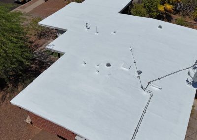 Aerial view of a flat white roof on a building, featuring various vents, pipes, and a satellite dish; surrounded by trees, rocks, and a paved area.