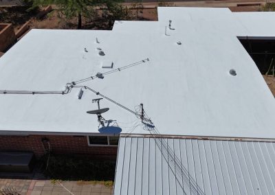 Aerial view of a flat, white-painted roof on a brick building with various rooftop equipment, including a satellite dish, vents, and cables, next to a corrugated metal patio roof and surrounding yard.