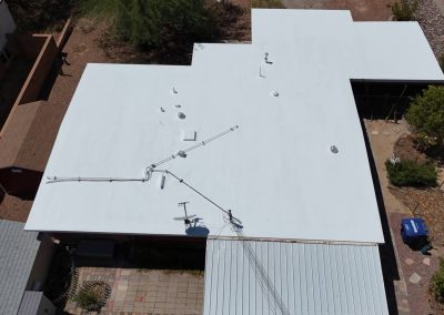 Aerial view of a flat white rooftop with various pipes, vents, and cables. The surrounding area includes a patio, vegetation, and a blue recycling bin near the corner of the house.