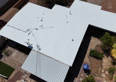 Aerial view of a white flat-roofed building with various vents and pipes, adjacent to a metal-roofed patio. Surrounding areas include paved walkways, brown landscaping, and some green plants and trees.
