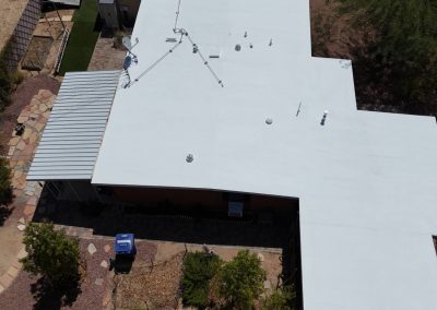 Aerial view of a building with a white flat roof, metal awning, and rooftop equipment, surrounded by a yard with trees, pathways, and a blue recycling bin near the entrance.