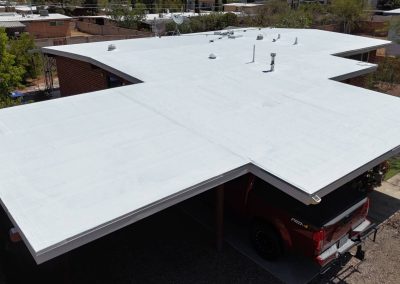 Aerial view of a modern flat-roofed building with a white coated surface, featuring several vents and pipes. A red pickup truck is parked in the carport beneath part of the roof. Trees and other houses are visible in the background.
