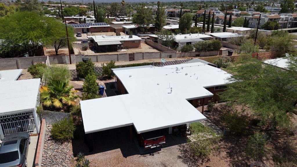 Aerial view of a residential neighborhood with flat-roofed homes, trees, and driveways. The main focus is a house with a bright white roof and a covered carport surrounded by desert landscaping.