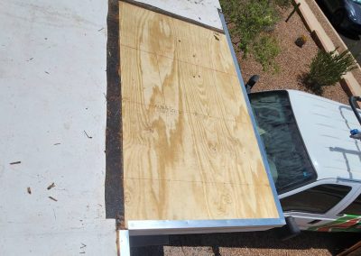 A section of a flat roof with new plywood installed, viewed from above. A white work van with ladders on top is parked next to the roof, and a landscaped yard with gravel and plants is visible below.