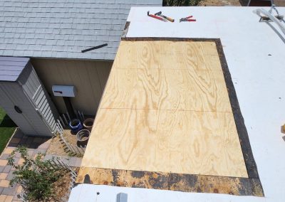 Plywood sheet partially installed on a flat rooftop, with tools and materials nearby. A shed, garden pots, and a lawn are visible next to the roof. The scene appears to show ongoing roof repair or construction work.