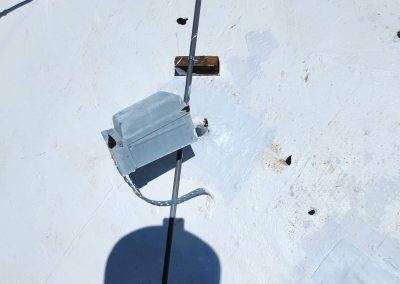 A metal electrical junction box with a conduit and cable is mounted on a white-painted surface, possibly a roof. There are patches and dark spots nearby, and a shadow from a round object appears in the foreground.