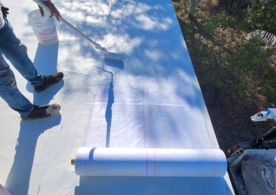 A person uses a paint roller to apply a white coating to a flat rooftop, with a roll of roofing material laid out beside them and a bucket of coating nearby. Trees and plants are visible in the background.