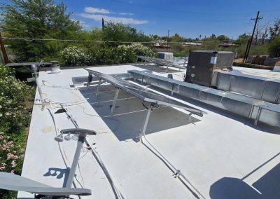 A flat white rooftop with metal ventilation ducts, solar panel mounts, an air conditioning unit, and a satellite dish, surrounded by trees and power lines under a partly cloudy sky.