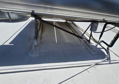 A solar water heater mounted on a flat white roof, supported by metal brackets, with visible rust and stains beneath the unit. Pipes and shadows can also be seen on the rooftop surface.