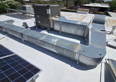 Rooftop view showing metal HVAC ductwork and a large ventilation unit next to solar panels, with other rooftops and green trees in the background under a clear sky.