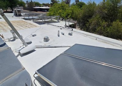 Flat white rooftop with various vents, skylights, and solar panels. Surrounding trees and a few nearby buildings are visible. The roof shows signs of weathering and has several mounted equipment and pipes.