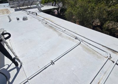 White rooftop with several metal pipes secured by brackets, running parallel to each other. Some trees and a partial view of a solar water heater are visible in the background.