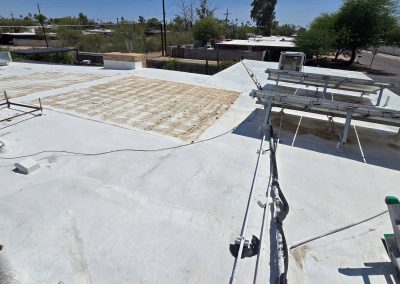 A flat white rooftop with pipe vents, some removed equipment leaving a rectangular patch, and metal racks for solar panels under a sunny sky, overlooking a residential area with trees and houses.