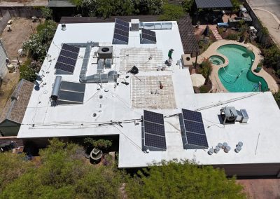 Aerial view of a white flat-roofed house with multiple solar panels installed, surrounded by trees, a swimming pool, and various outdoor furniture in the backyard. Two people are working on the roof.