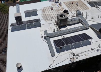 Aerial view of a flat white rooftop with solar panels, HVAC units, ductwork, and scattered equipment. Some roof sections appear newly covered, while others show exposed older material. Trees border the rooftop.