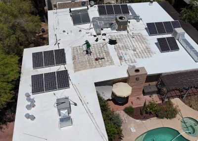 Aerial view of a flat white rooftop with multiple solar panels, HVAC units, and a person working on the roof. Below, there is a backyard with a swimming pool, patio furniture, trees, and plants.
