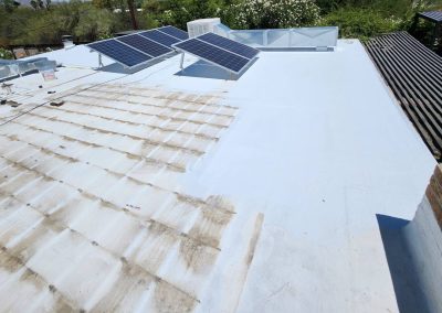 Flat white rooftop with solar panels and HVAC units. The roof shows a section with some dirt or staining and clean areas. Nearby, there are green trees, shrubs, and a shaded patio with slatted cover.