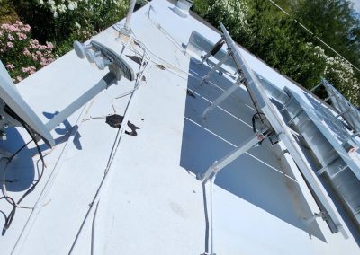 View of a white rooftop with solar panel mounts and electrical cables, surrounded by greenery and flowers in the background under a blue sky.