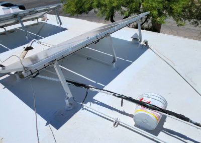 Two tilted solar panels mounted on a flat white rooftop, with wiring and a white bucket nearby. Trees and a street with parked cars are visible in the background.