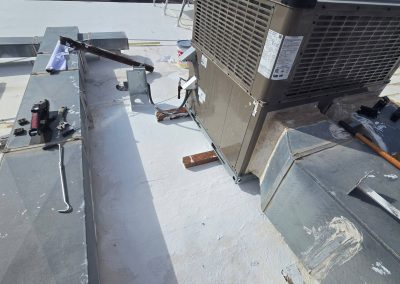 Metal HVAC ductwork and tools are scattered on a flat white rooftop near a large air conditioning unit, with various equipment and materials visible in the background.