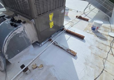 An air conditioning unit is mounted on a flat rooftop near solar panels, surrounded by wooden planks, cables, and paint buckets. The roof appears to be in the process of being painted or repaired.