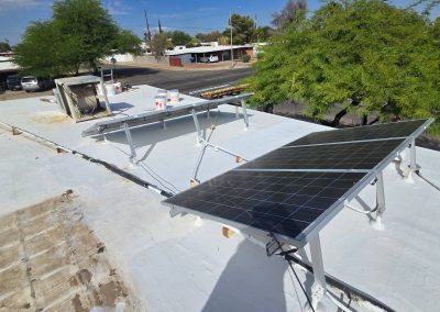 Two solar panels installed on a flat white rooftop, surrounded by trees and nearby residential buildings under a partly cloudy sky. A ventilation unit and tools are also visible on the roof.