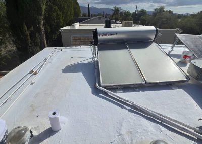 A rooftop with a solar water heater system, solar panels, pipes, a can of paint, and a paper towel roll under a partly cloudy sky with trees and mountains in the background.