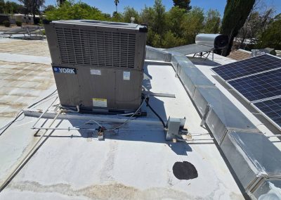 A rooftop HVAC unit labeled "YORK" is installed near metal ductwork and solar panels. The roof surface is white with some patches, and trees are visible in the background under a clear sky.