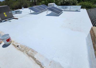 A flat white rooftop with solar panels, air conditioning units, paint buckets, and a partially visible person standing on the left side. Bushes and trees are seen in the background.