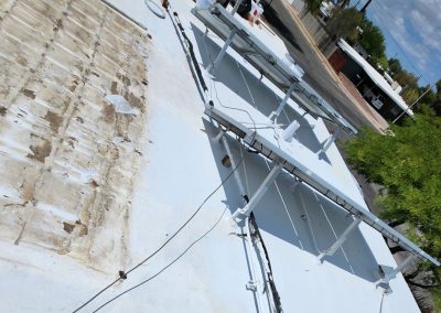 View of a white-painted rooftop with mounted solar panels and some exposed, weathered sections. Nearby, there are trees and a street with parked cars and buildings in the background.