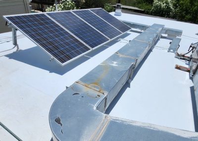 Two solar panels are mounted on a flat white rooftop near a large metal HVAC duct system, with greenery visible in the background.