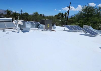 A flat white rooftop with solar panels, HVAC units, and other equipment under a clear blue sky, surrounded by trees and distant mountains.